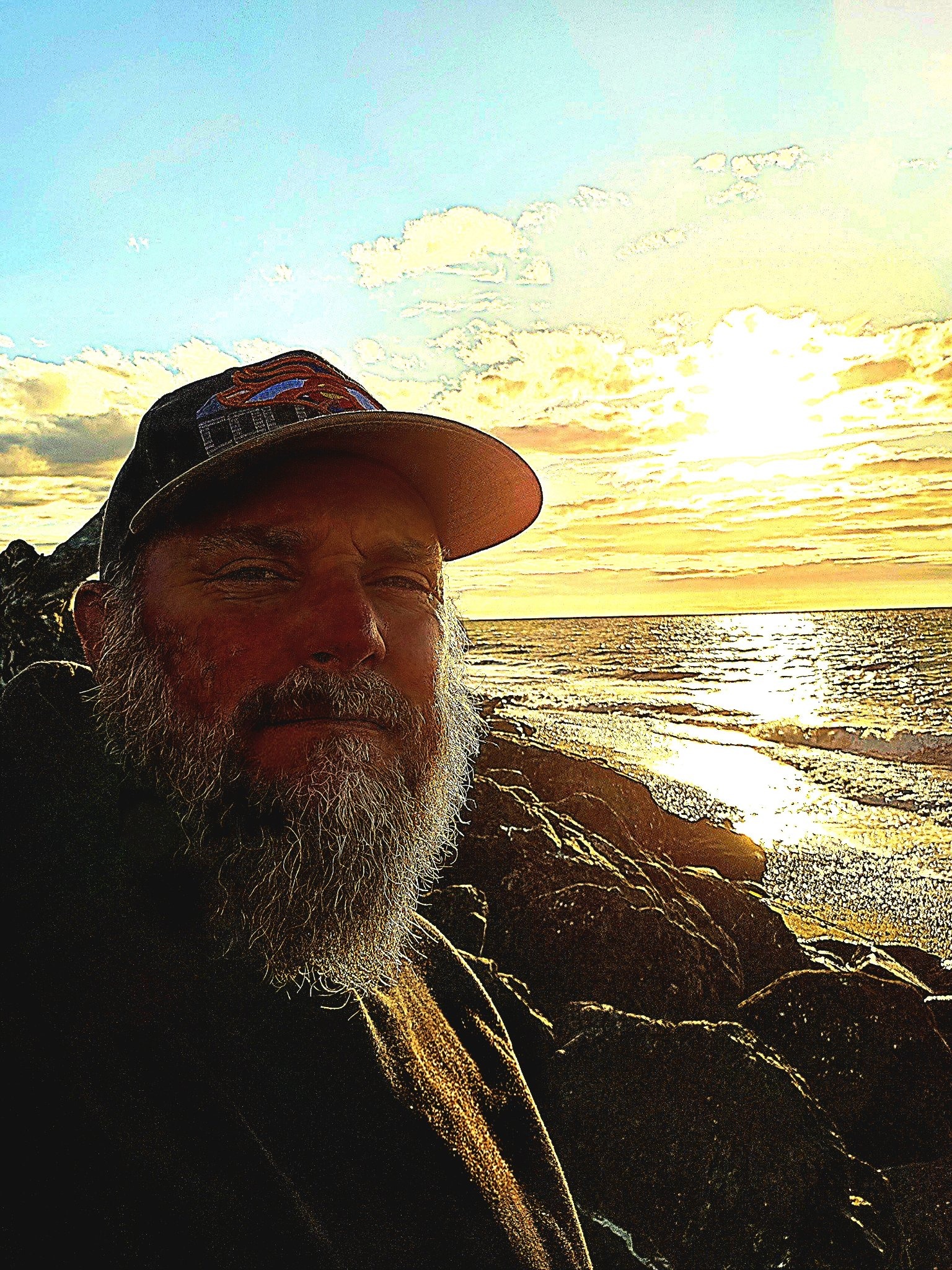 Anthony (Tony) in a cap at sunset on a rocky beach, the ocean and evening sky behind him